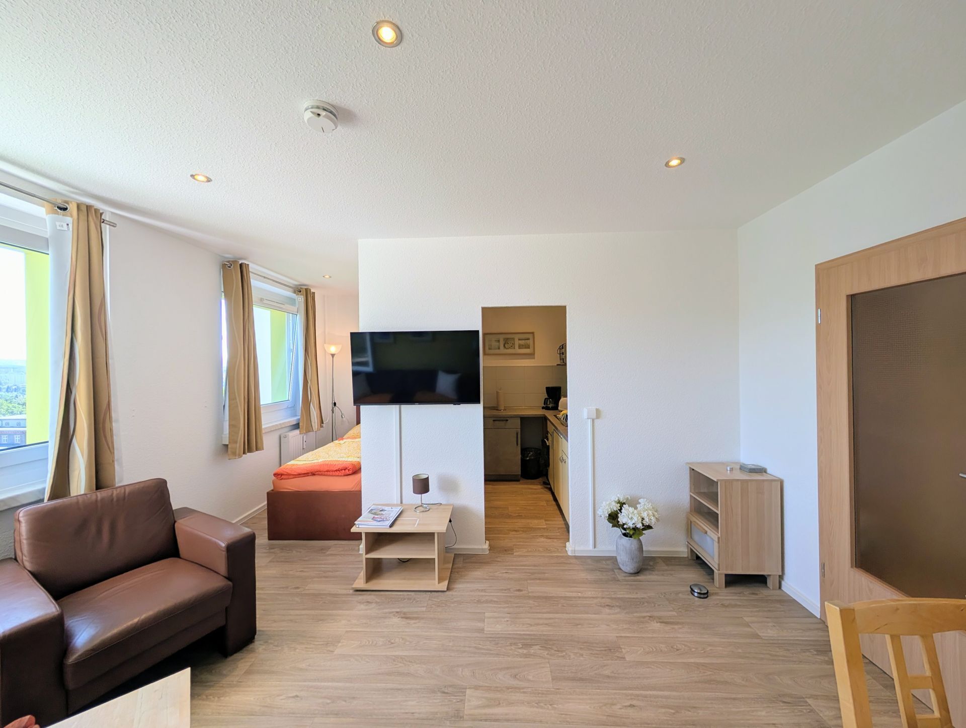 Wohnzimmer mit Blick Richtung Küche und Schlafbereich inklusive Sessel, weiterem Schrank samt Deko und kleinem Schrank unter hängendem Fernseher