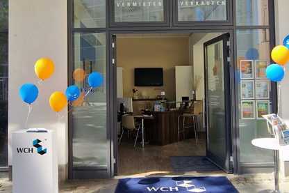 Innenstadtbüro mit offener Glastür, blau-gelben Luftballons und "WCH"-Logo auf Säulen und Fußmatte