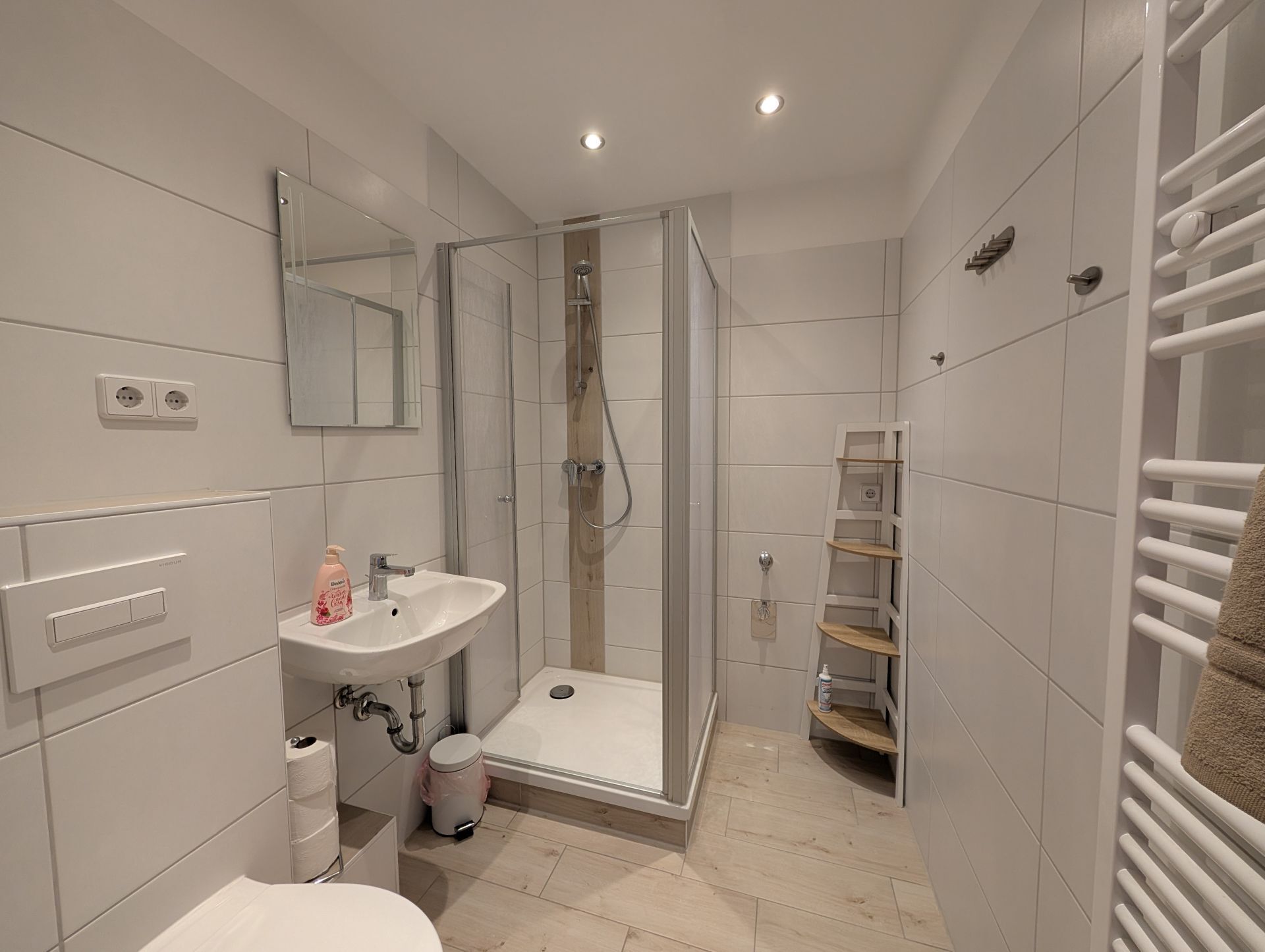 Badezimmer mit Dusche, LED-Deckenspots, Handtucheizkörper, WC und Waschbecken mit Seifenspender sowie Badspiegel und Badregal