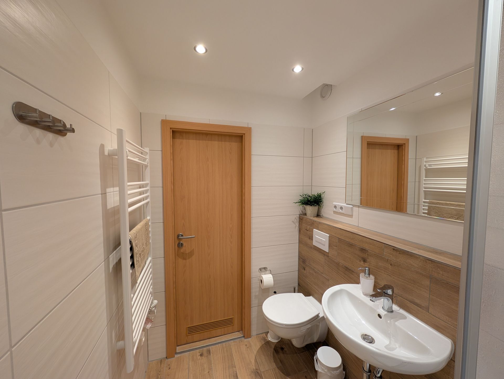 Badezimmer mit Blickrichtung Badtür, LED-Deckenspots, Handtucheizkörper, WC und Waschbecken mit Seifenspender sowie Badspiegel und Badregal