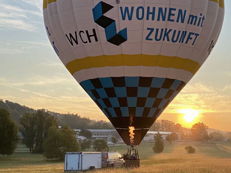 Heißluftballon mit Werbung "WCH Wohnen mit Zukunft" beim Start in einem Feld bei Sonnenuntergang.