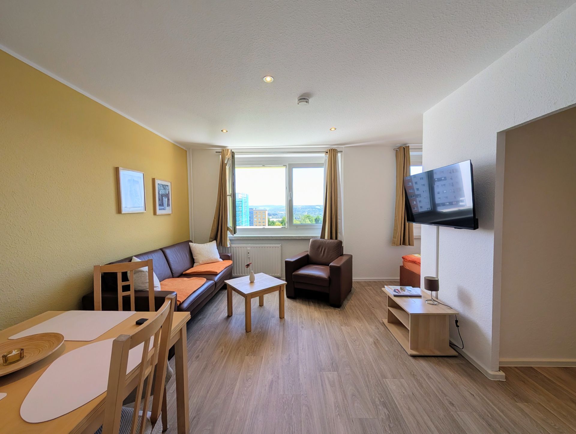 Wohnzimmer mit Blick Richtung geöffnetem Fenster inklusive Sofa, Couchtisch, Sessel, Essecke und kleinem Schrank unter hängendem Fernseher