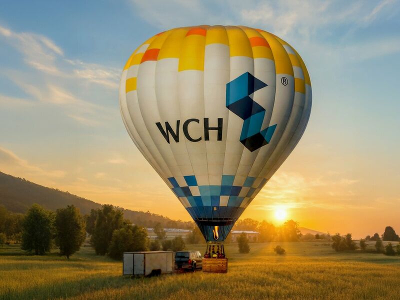 Heißluftballon mit Werbung "WCH Wohnen mit Zukunft" beim Start in einem Feld bei Sonnenuntergang.