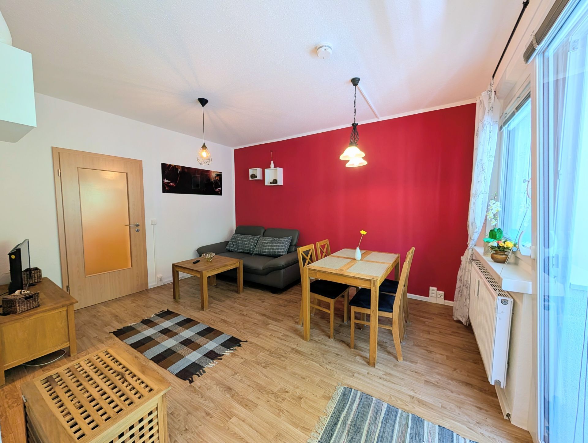 voll eingerichtetes Wohnzimmer inklusive Sofa, Couchtisch, Essbereich, Fernseher auf Lowboard sowie rechtsseitig gekippter Balkontür und Blick auf die rot gemalerte Wand hinterm Sofa