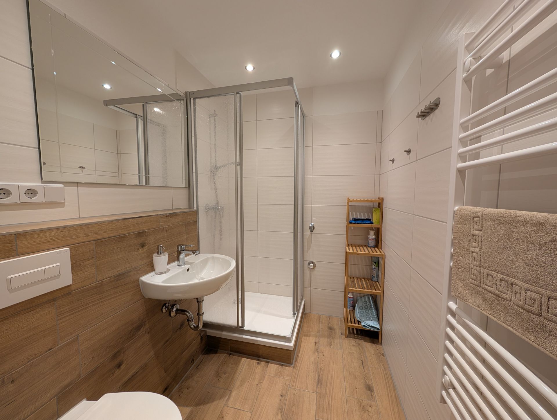 Badezimmer mit Dusche, LED-Deckenspots, Handtucheizkörper, WC und Waschbecken mit Seifenspender sowie Badspiegel und Badregal