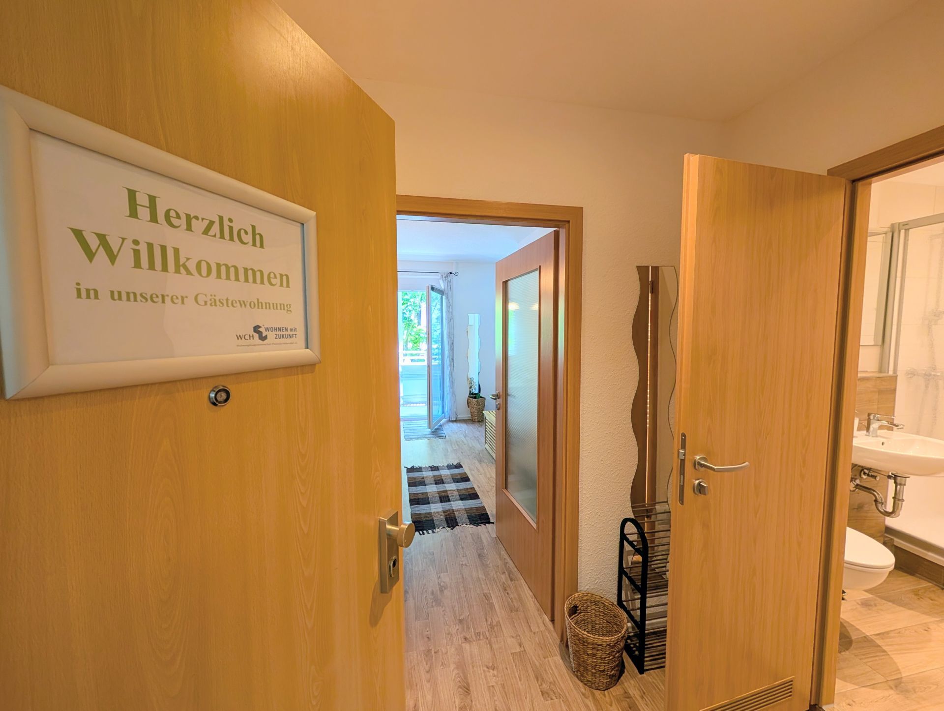 geöffnete Wohnungstür mit Herzlich-Willkommen-Schild, die sowohl einen Blick in den Flur als auch ins Badezimmer und ins Wohnzimmer erlaubt