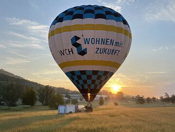 Heißluftballon mit Werbung "WCH Wohnen mit Zukunft" beim Start in einem Feld bei Sonnenuntergang.