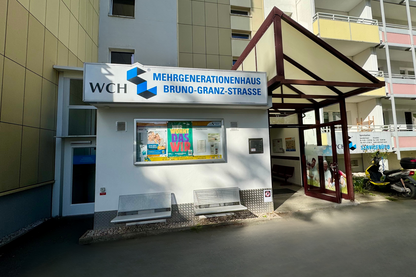 Kleines weißes Gebäude mit Vordach, Schild "Mehrgenerationenhaus Bruno-Granz-Straße" und "WCH"-Logo, neben einem größeren Wohnkomplex