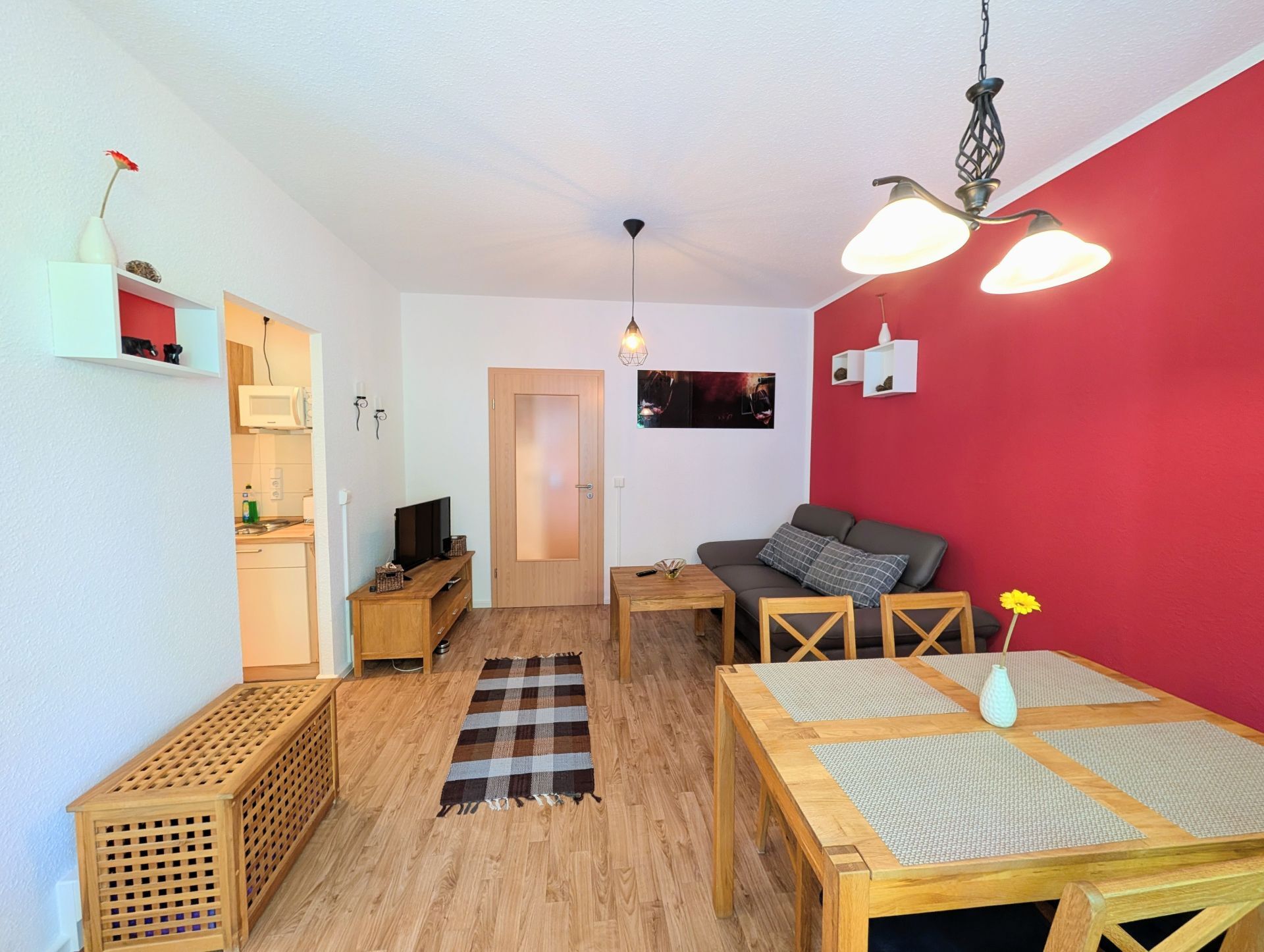 voll eingerichtetes Wohnzimmer inklusive Sofa, Couchtisch, Essbereich, Fernseher auf Lowboard sowie Blick auf geschlossene Wohnzimmertür und linksseitig Blick in die Küche