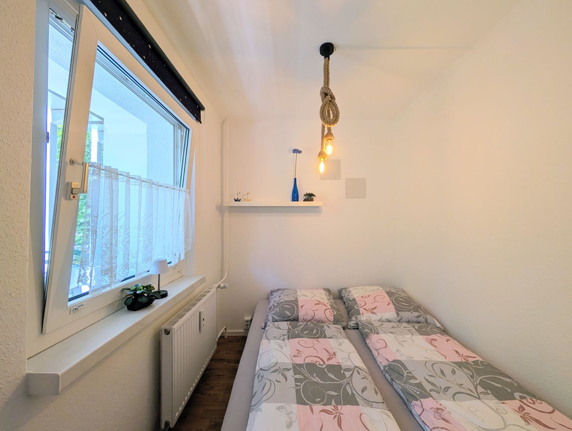 Schlafbereich mit Bett inklusive frisch bezogener Bettwäsche, einer Deckenlampe und links einem gekipptem Fenster mit Deko auf dem Fensterbrett