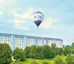 Heißluftballon mit "WCH" Logo über einem Wohngebäude in grüner Umgebung.