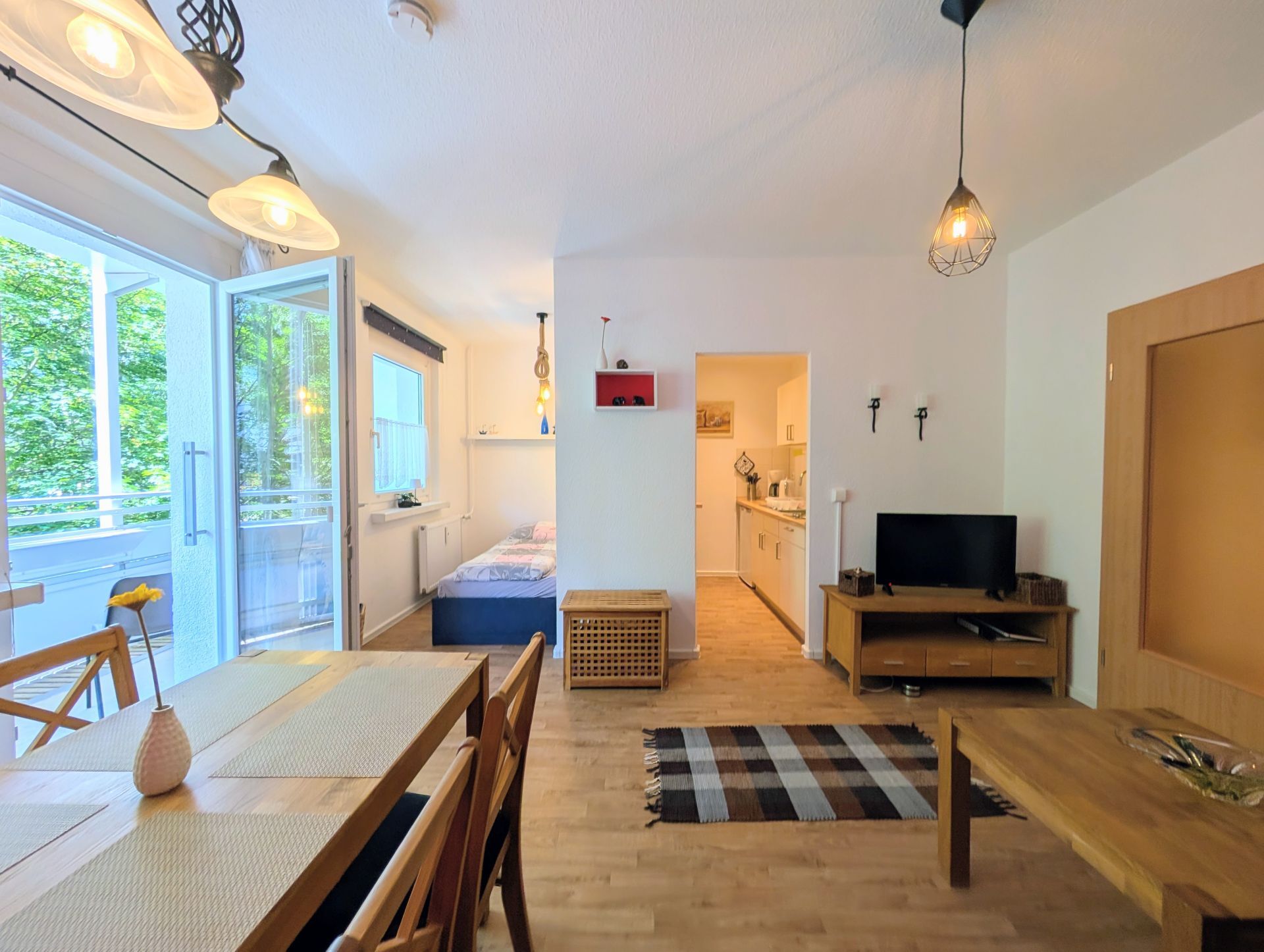 voll eingerichtetes Wohnzimmer inklusive Couchtisch, Essbereich, Fernseher mit Lowboard sowie linksseitig geöffneter Balkontür und Blick auf Küche und Schlafbereich
